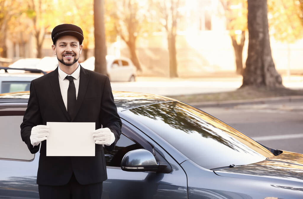 Handsome chauffeur with blank paper sheet near luxury car