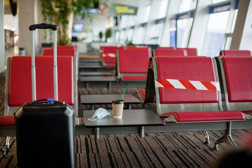 A medical mask and a cup of coffee sit on a table in an empty airport, next to the red seats. Coronavirus, social distance, isolation.