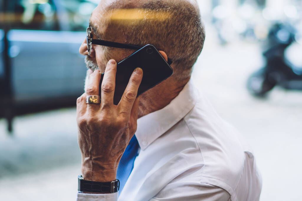 Side view of aged male in white shirt with grey short hair sitting and talking on smartphone against background of city street and road