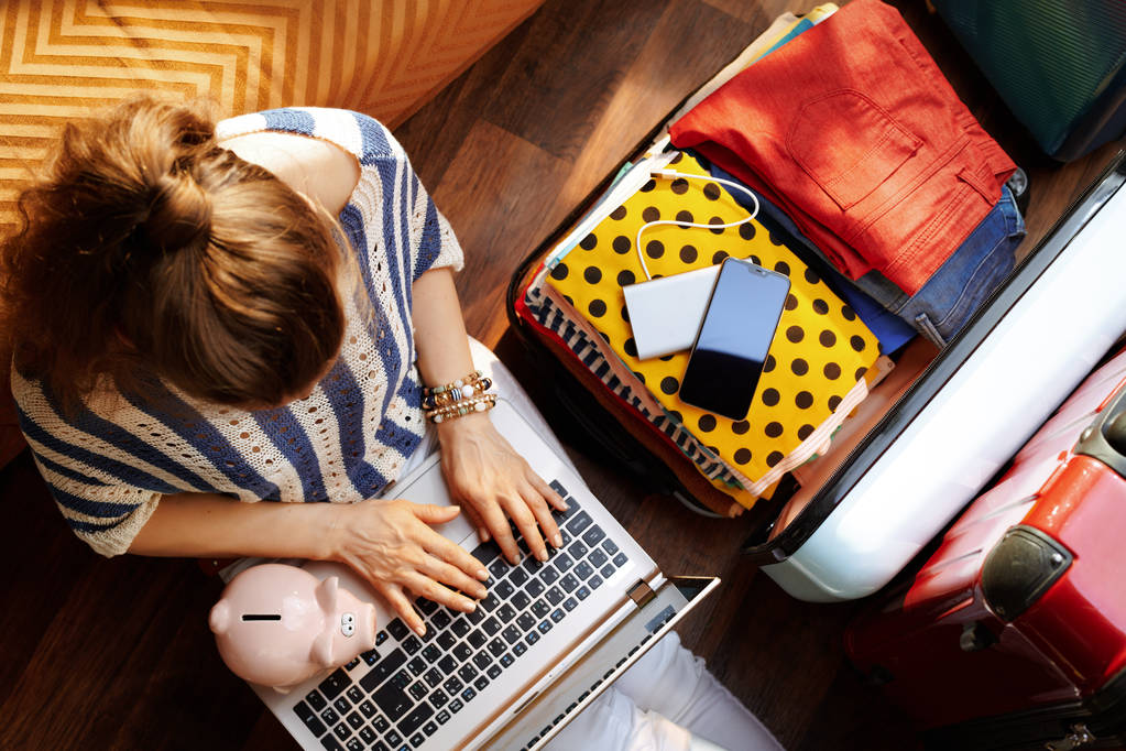 Upper view of stylish woman in white pants and striped blouse with piggy bank near open travel suitcase booking economy hotel online at modern home in sunny summer day.