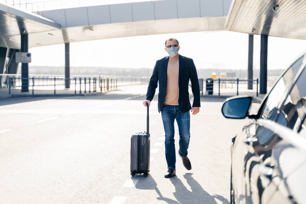 Business traveler pulls suitcase from modern airport terminal, walks to car, wears medical disposable mask on face, prevents from coronavirus during dangerous pandemic situation. Full length shot