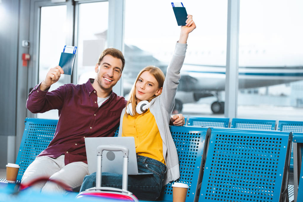 happy girlfriend holding passport above head near smiling boyfriend in airport near luggage